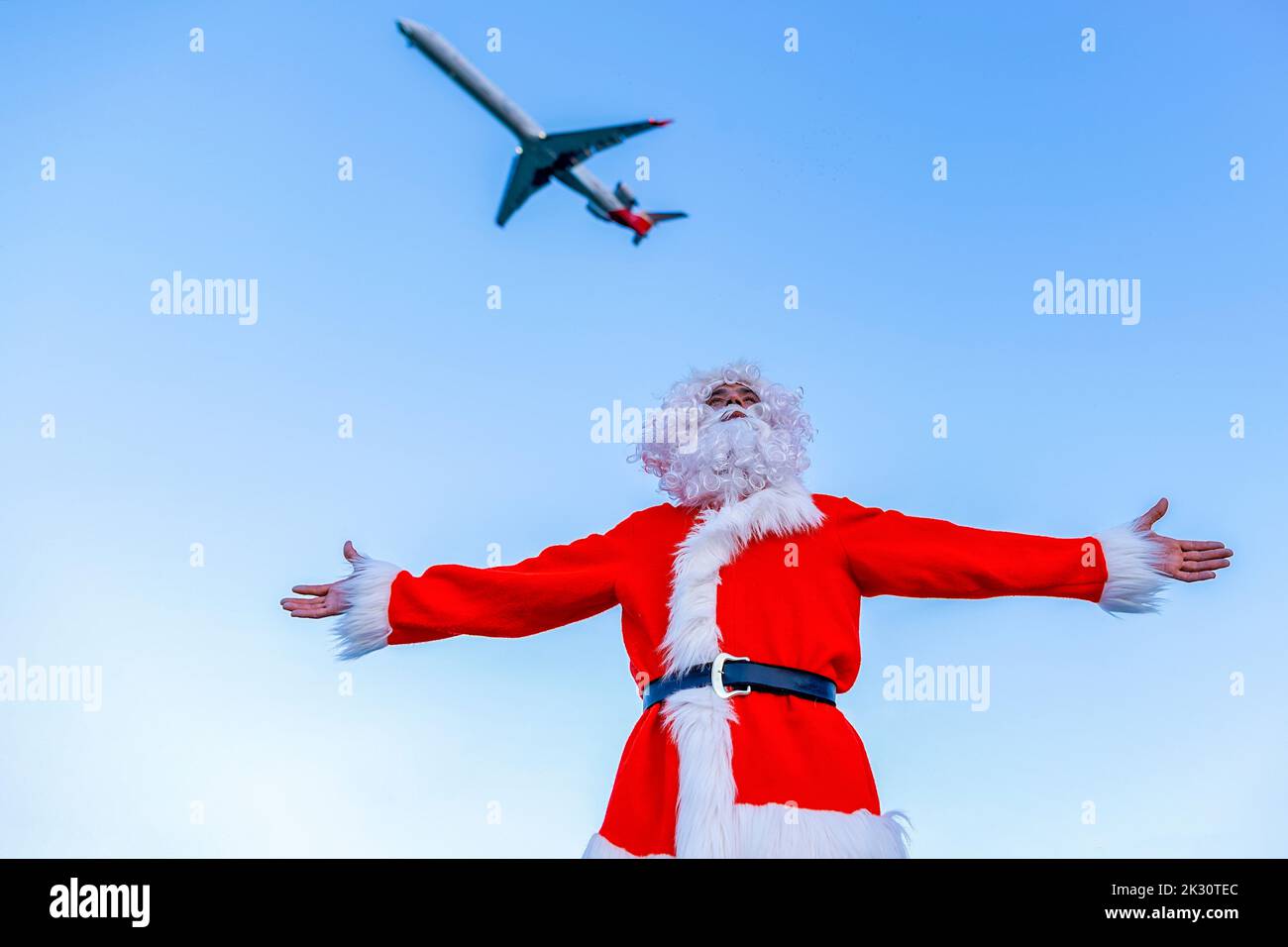 Man wearing Santa Claus costume standing with arms outstretched below flying airplane Stock Photo