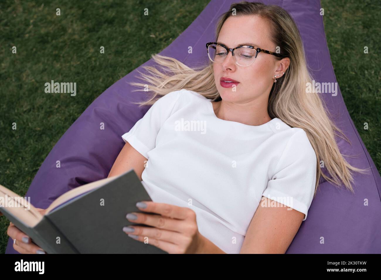 Businesswoman wearing eyeglasses reading book lying on bean bag Stock