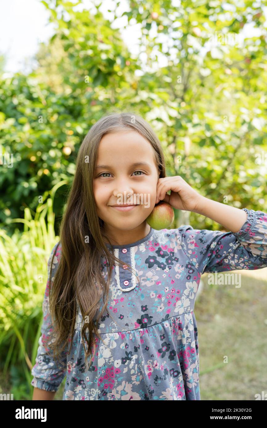 Smiling girl holding apple near ear in garden Stock Photo