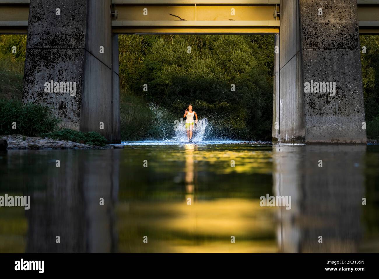 Active young woman running through river water under bridge Stock Photo
