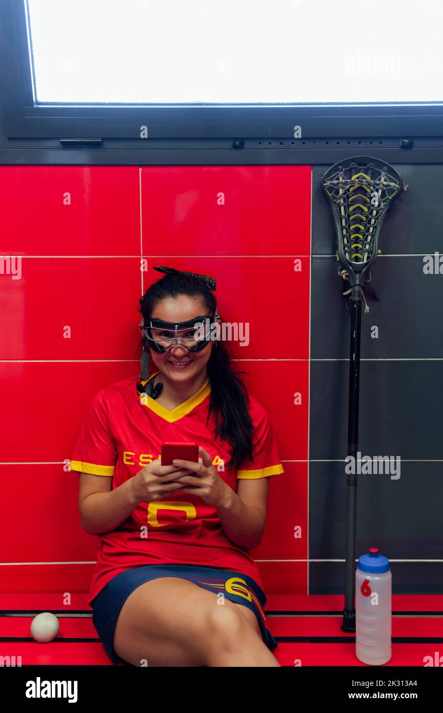 Happy woman wearing protective eyewear sitting with mobile phone in locker room Stock Photo