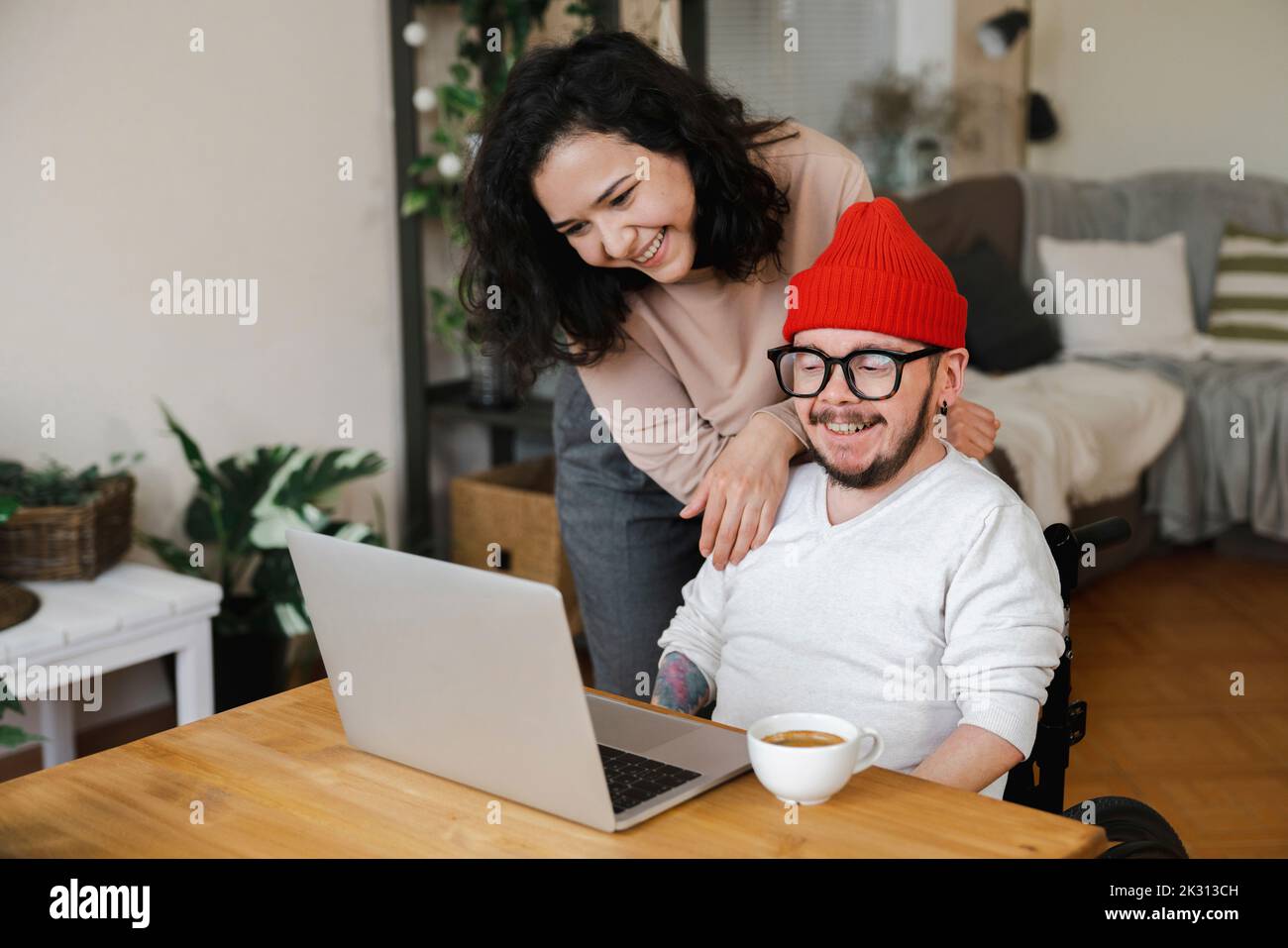 Smiling woman standing by boyfriend in wheelchair looking at laptop Stock Photo