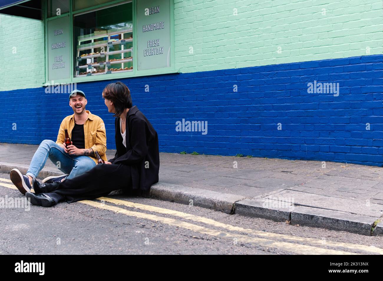 Happy man holding beer bottle sitting with boyfriend on sidewalk Stock Photo