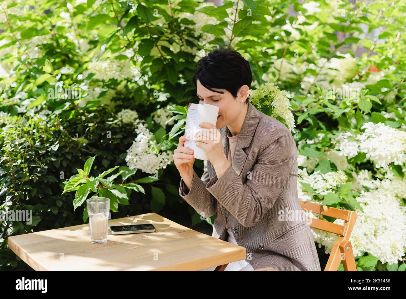 Businesswoman covering face with book sitting at table by plants Stock Photo