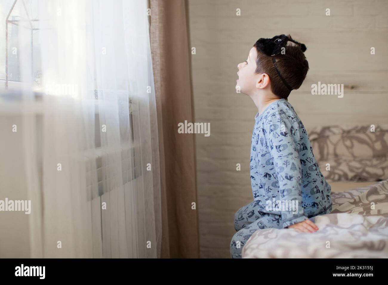 Boy with sleep mask looking through window sitting on bed Stock Photo