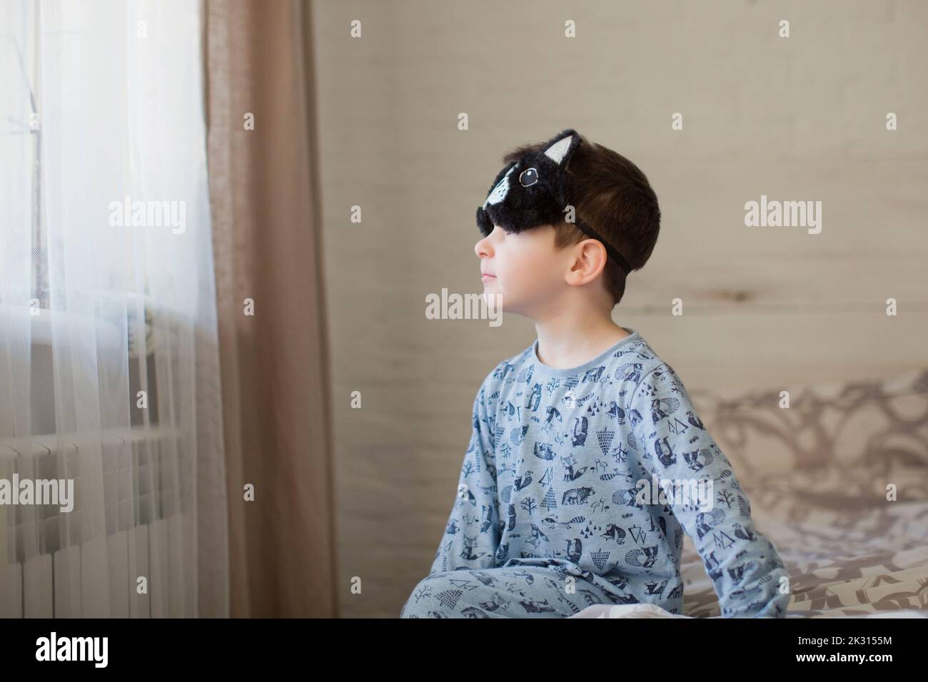 Cute boy with sleep mask looking through window sitting on bed Stock Photo