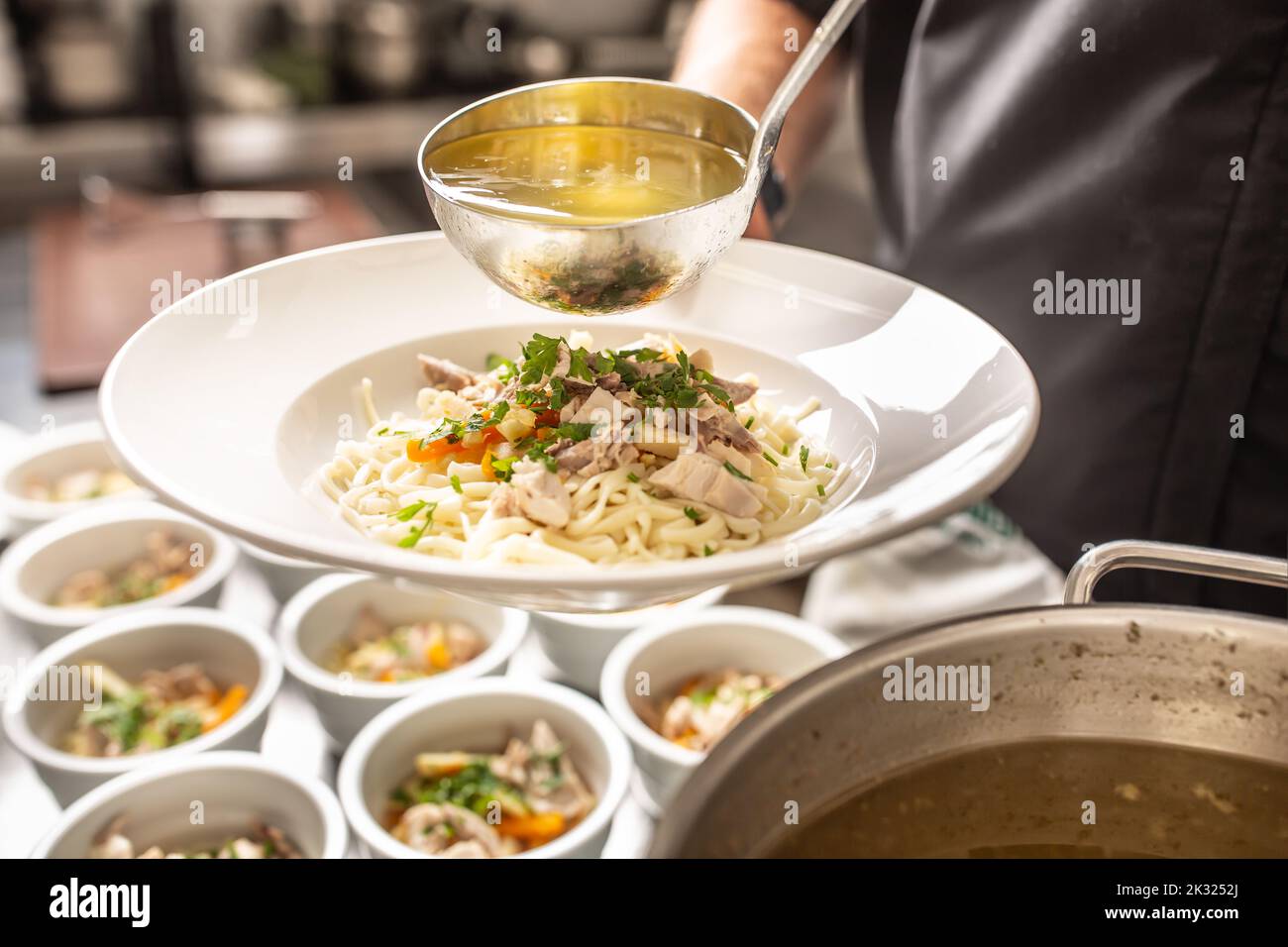 Chef in restaurant kitchen pours broth into a chicken soup with noodles, meat and vegetable. Stock Photo