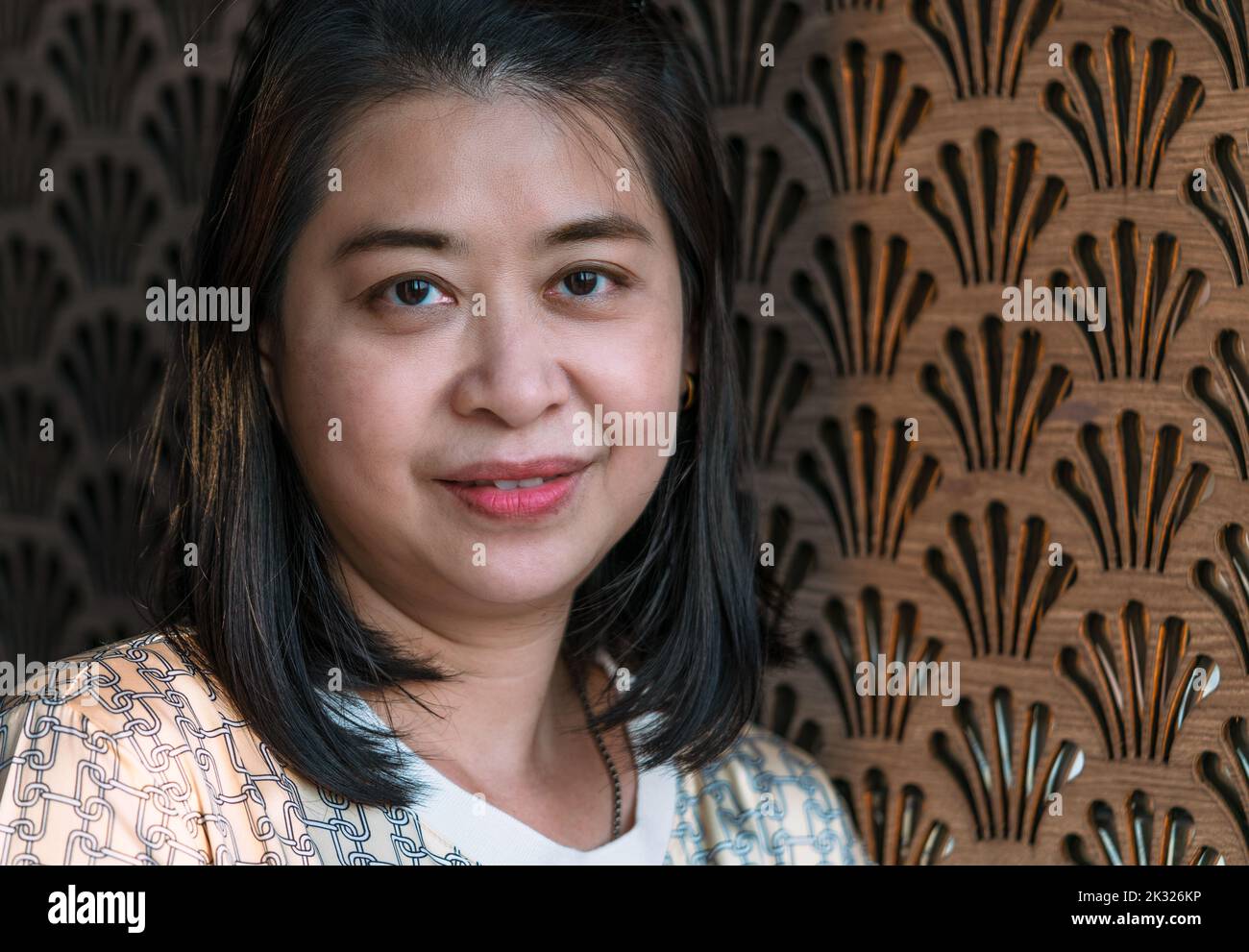 Portrait of beautiful Asian middle-aged woman with brown wooden wall, smiling face, natural sunlight, blank space for text and design. Stock Photo