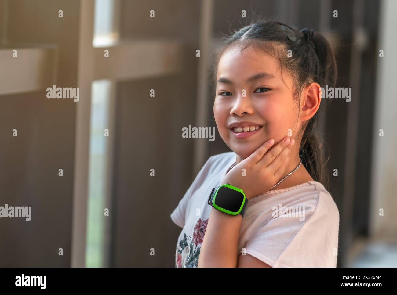 Portrait of cute Asian child girl, standing next to glass window, smiling and looking at camera, cute pose with one hand touching at cheek, wearing di Stock Photo