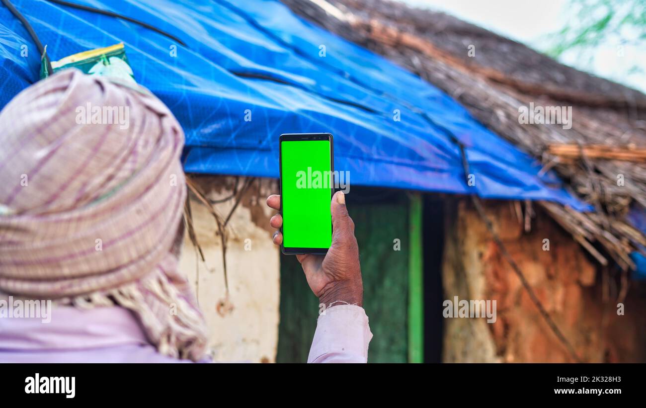Old Indian man taking selfie and showing a green screen cell phone with rural village life concept background. Stock Photo