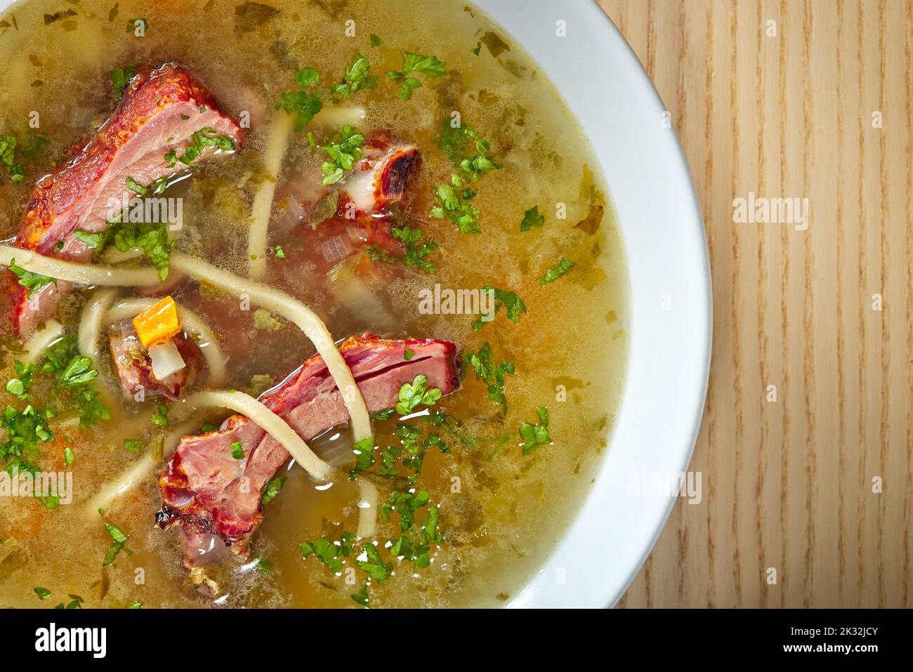 Soup of smoked ribs with vegetables and herbs in a white bowl standing on wooden background - top view Stock Photo