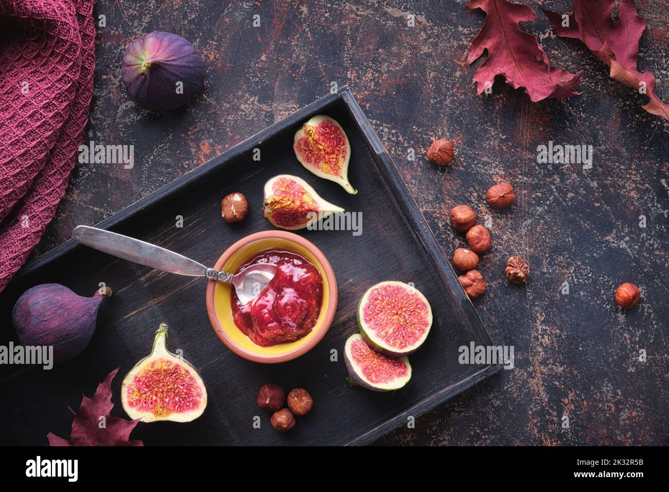 Autumntime background with fig jam, spoon, nuts and fresh halved fig fruits on black wooden tray. Dark brown background with dry Fall leaves. Stock Photo