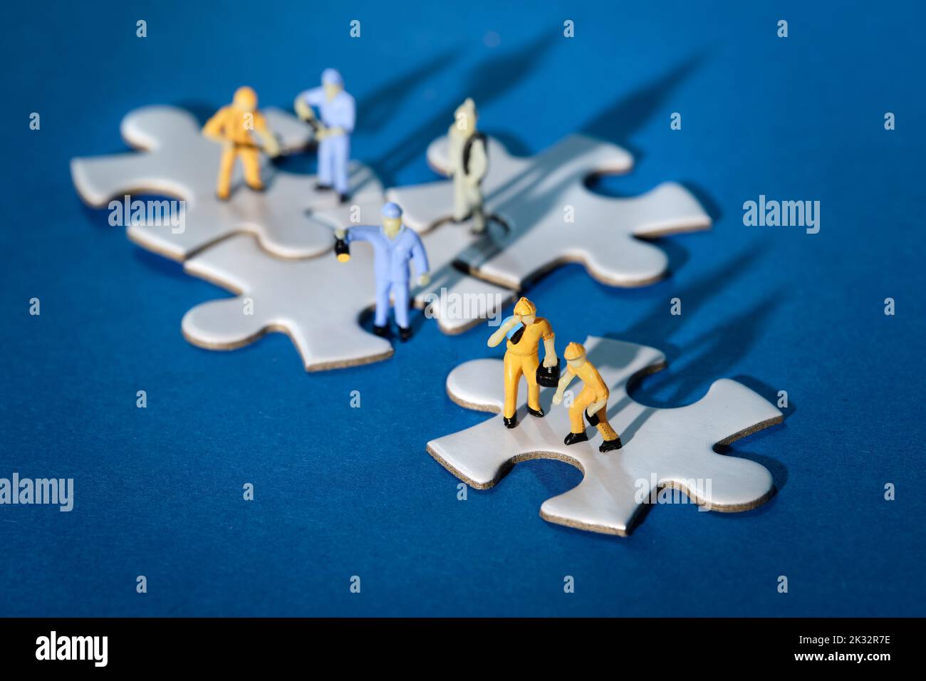 Team of tiny worker miniature figures on linked jigsaw puzzle pieces island on blue paper. Dramatic light with long shadows. Stock Photo