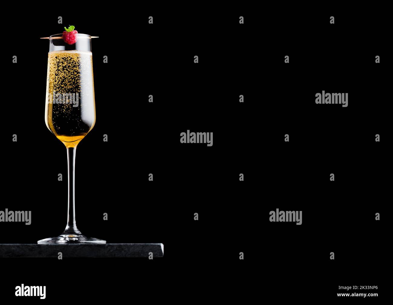 Elegant glass of yellow champagne with rasspbery on stick on black marble board on black background. Stock Photo