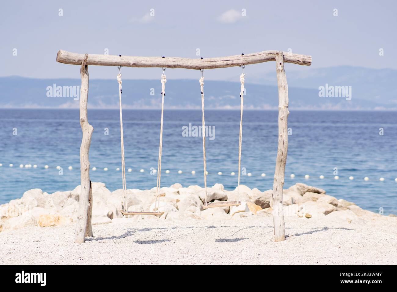 A pair of wooden swings tied with ropes at the beach against the background view of the sea. Stock Photo