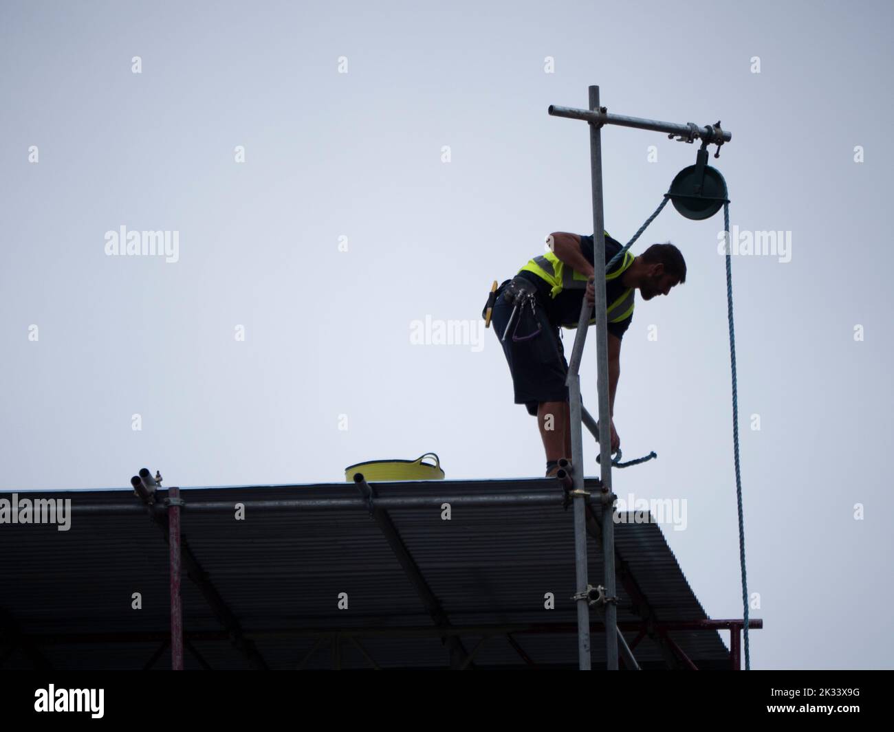 builder, construction worker roofer, standing on rooftop  scaffolding tower, with rope and pulley hoisting up materials for building work Stock Photo