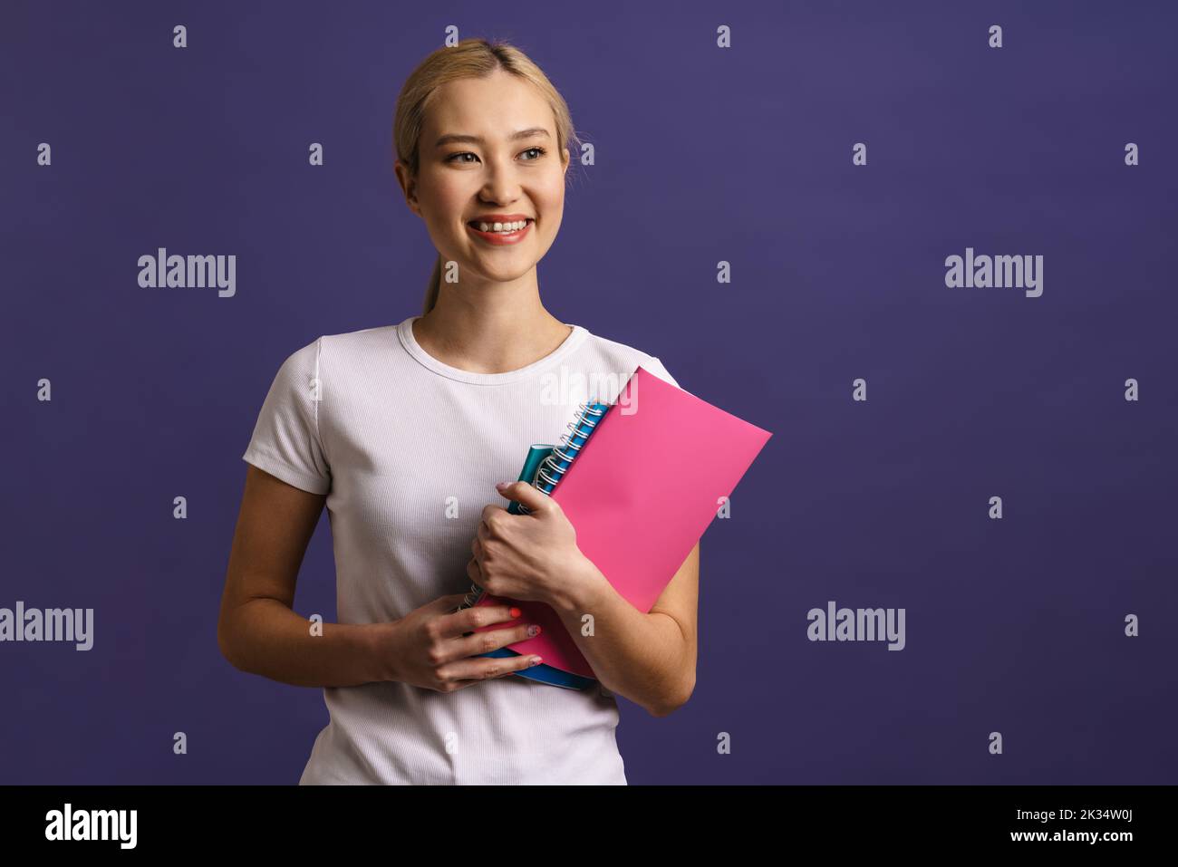 Smiling young blonde asian woman standing isolated over blue background carrying textbooks Stock Photo