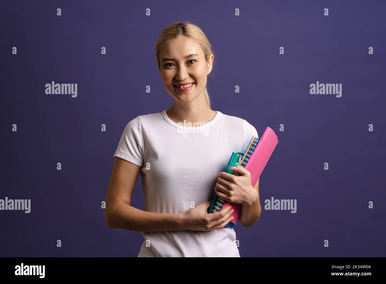 Smiling young blonde asian woman standing isolated over blue background carrying textbooks Stock Photo