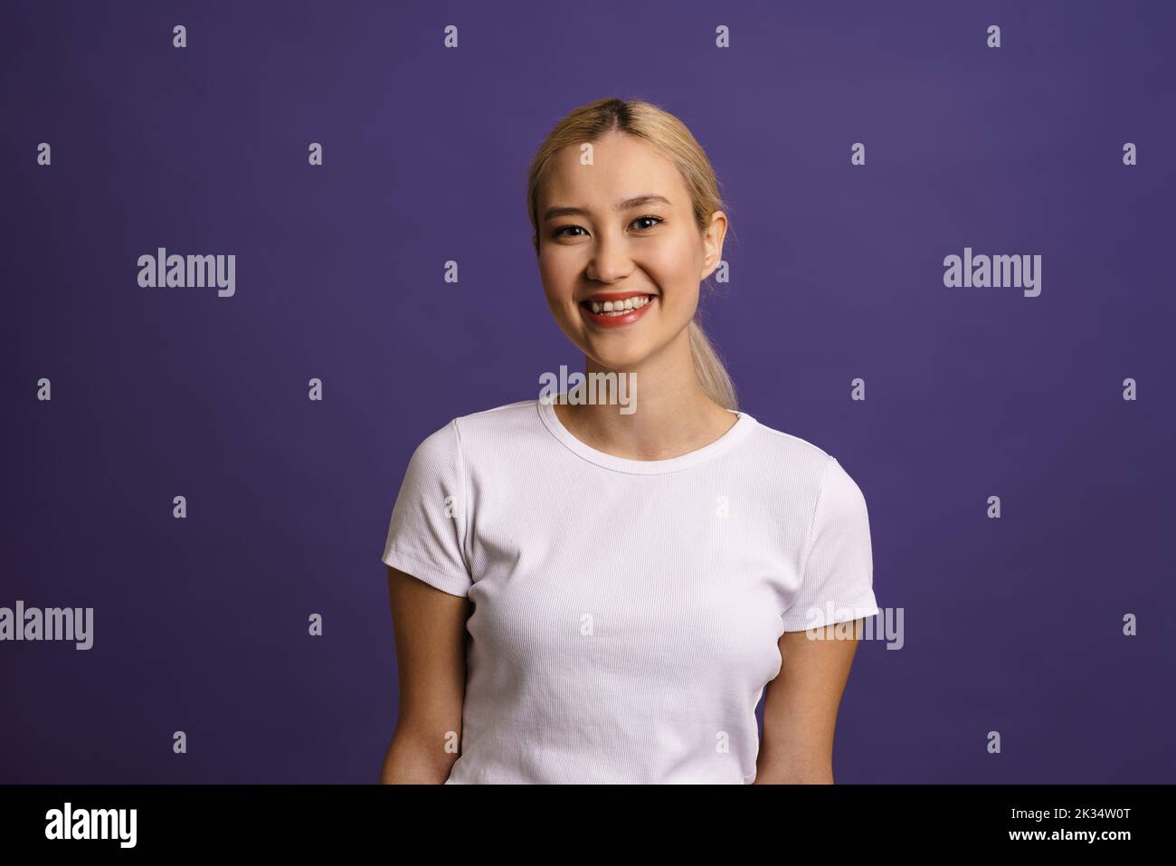 Smiling young blonde asian woman standing isolated over blue background looking at camera Stock Photo