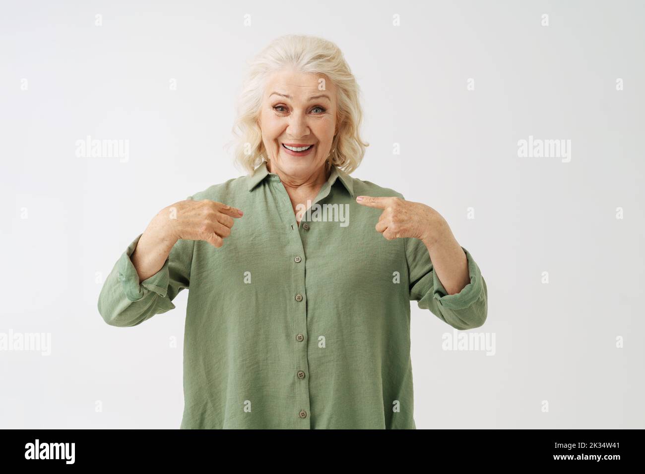 Grey senior woman in shirt smiling while pointing fingers at herself isolated over white background Stock Photo