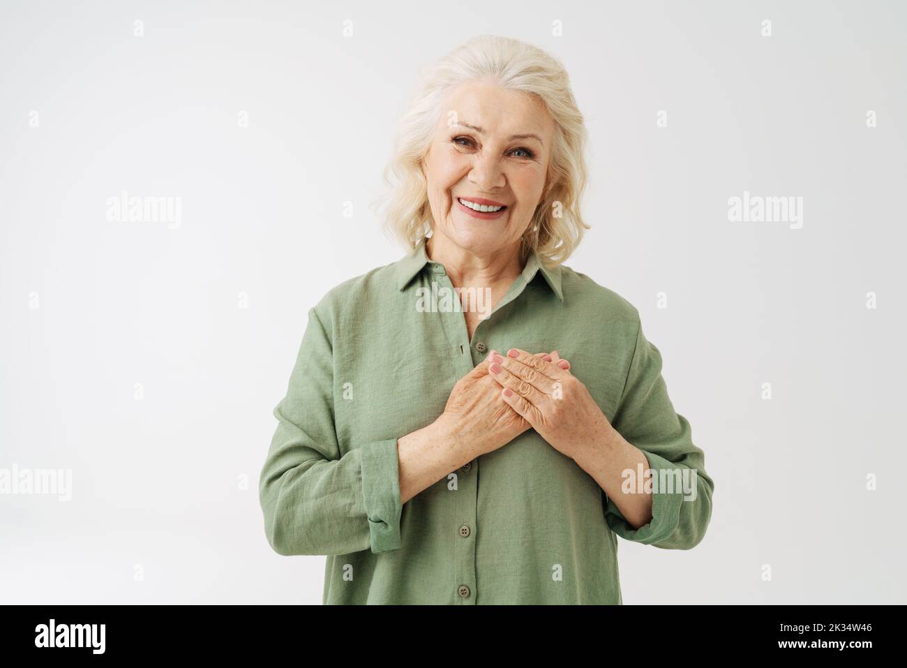 Grey senior woman in shirt smiling while holding her hands on chest isolated over white background Stock Photo