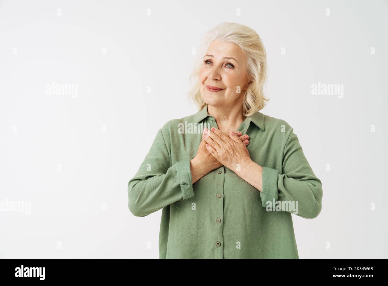 Grey senior woman in shirt smiling while holding her hands on chest isolated over white background Stock Photo