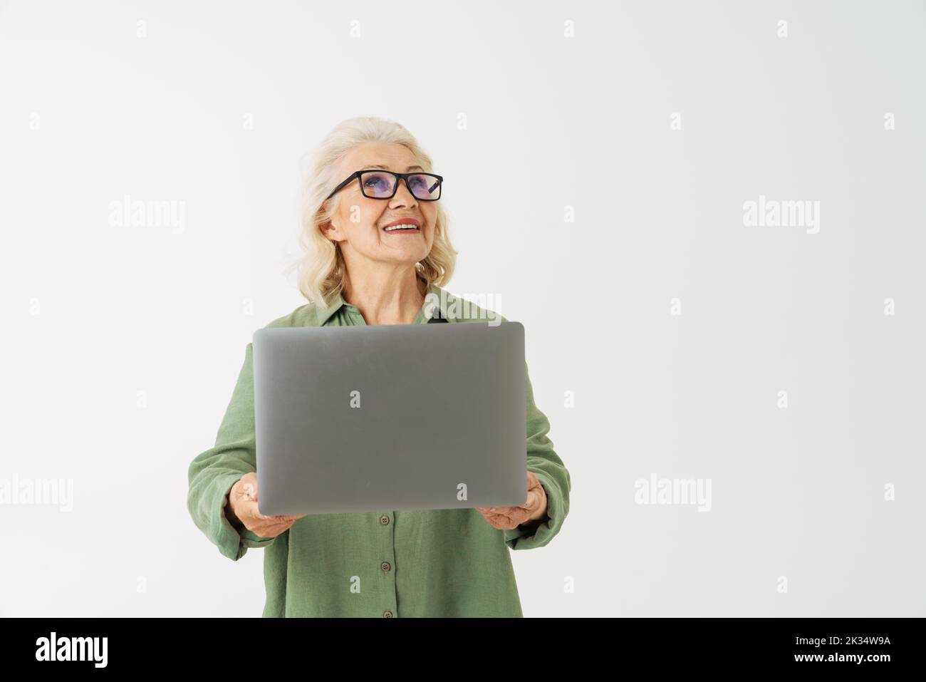 Grey senior woman in eyeglasses smiling while posing with laptop isolated over white background Stock Photo