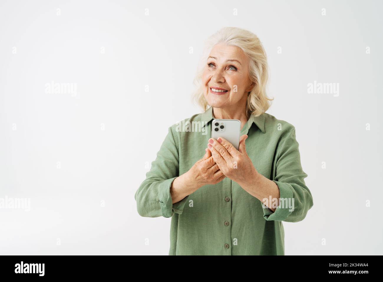 Grey senior woman in shirt smiling while using mobile phone isolated over white background Stock Photo