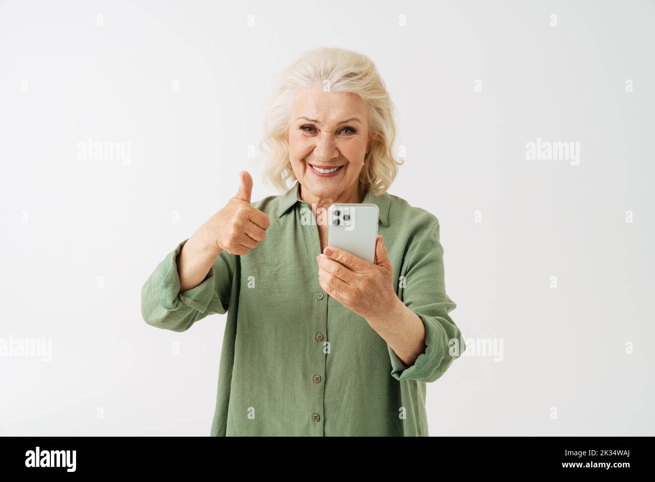 Grey senior woman showing thumb up while using mobile phone isolated over white background Stock Photo