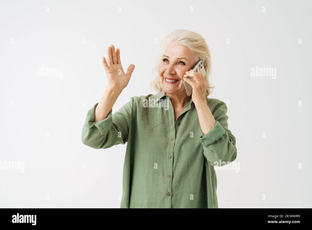 Grey senior woman in shirt gesturing while talking on mobile phone isolated over white background Stock Photo