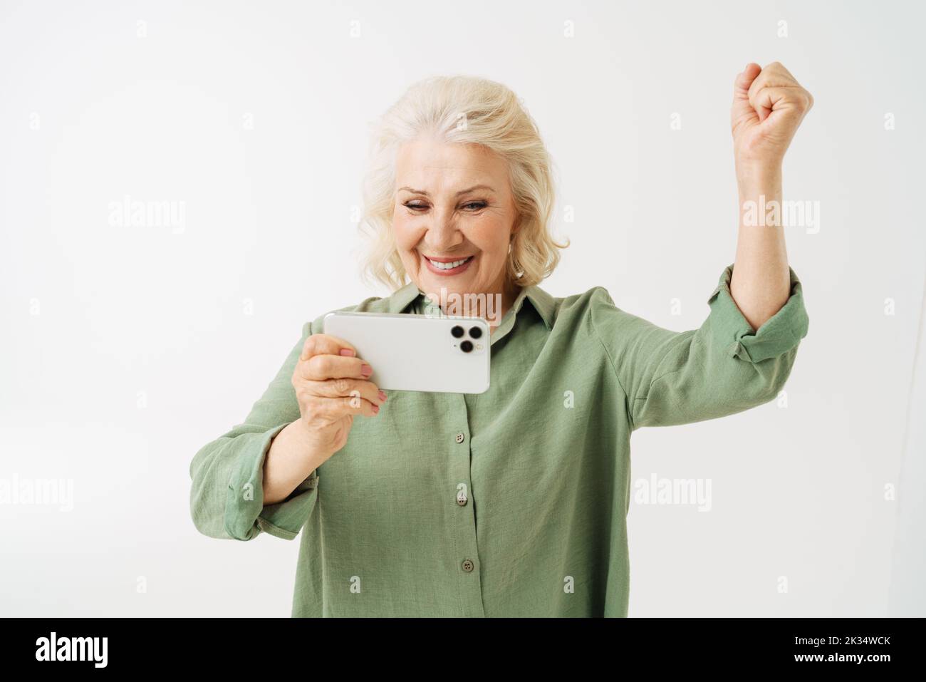 Grey senior woman gesturing while playing online game on cellphone isolated over white background Stock Photo