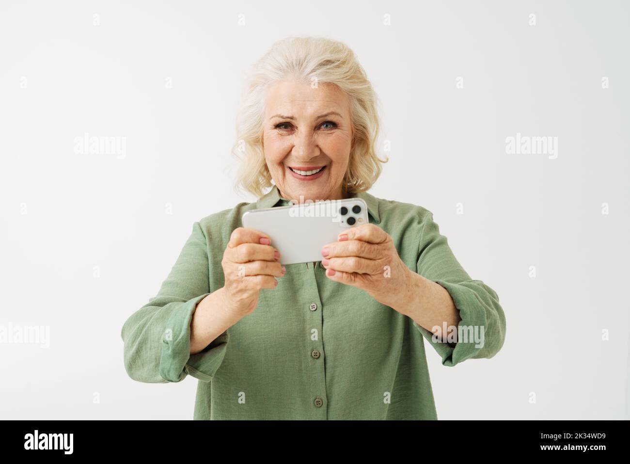 Grey senior woman smiling while playing online game on mobile phone isolated over white background Stock Photo