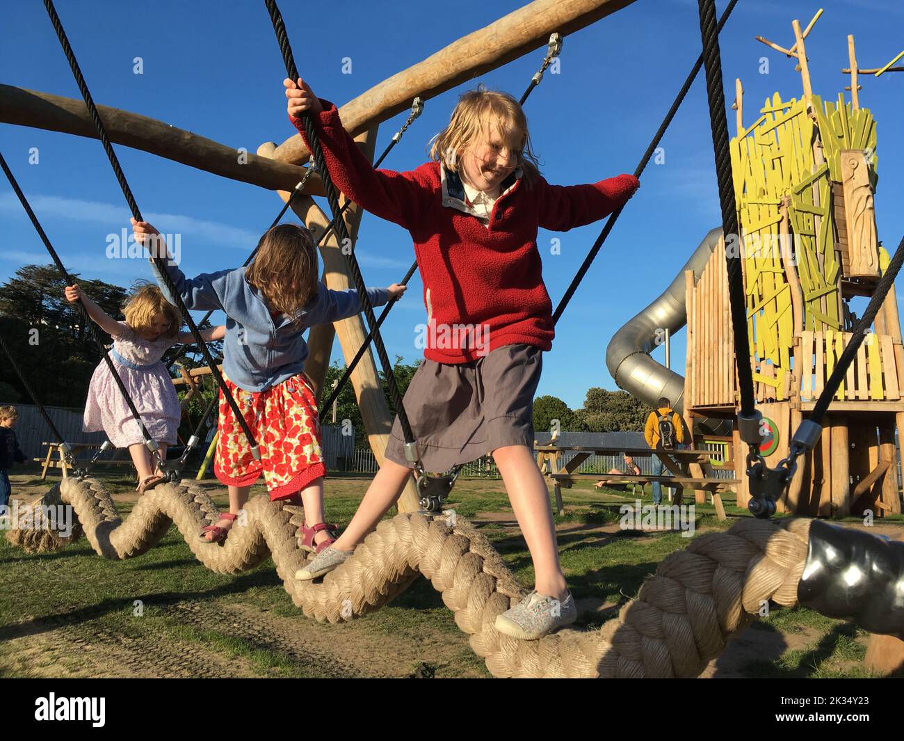 Three young girls / children / child / kids / kid play in the playground on the day with sun and blue sky. The play equipment involves a very thick climbing rope suspended horizontally, from a frame. The 3 girls ARE model released. (132) Stock Photo