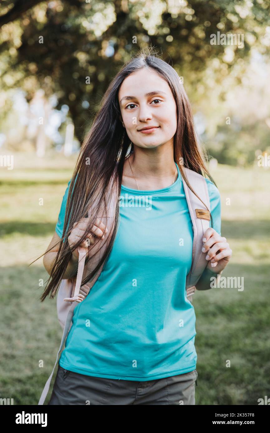 Young student girl, carrying a backpack and looking at the camera with a serene smile. Confidence looking  Stock Photo