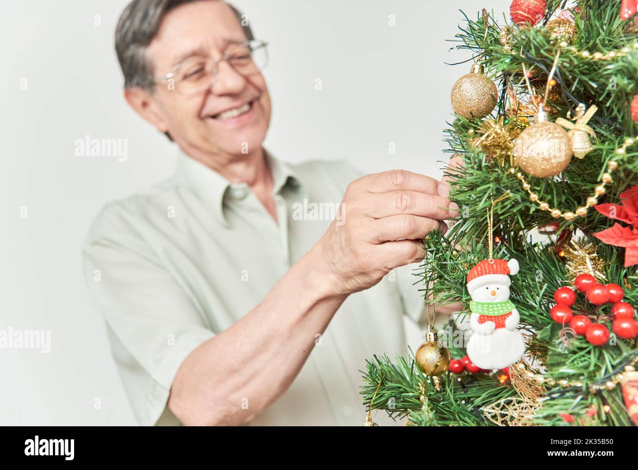 Happy senior latin man smiling while decorating a traditional Christmas tree at home. Selective focus on the foreground, the hands hanging a bauble fr Stock Photo