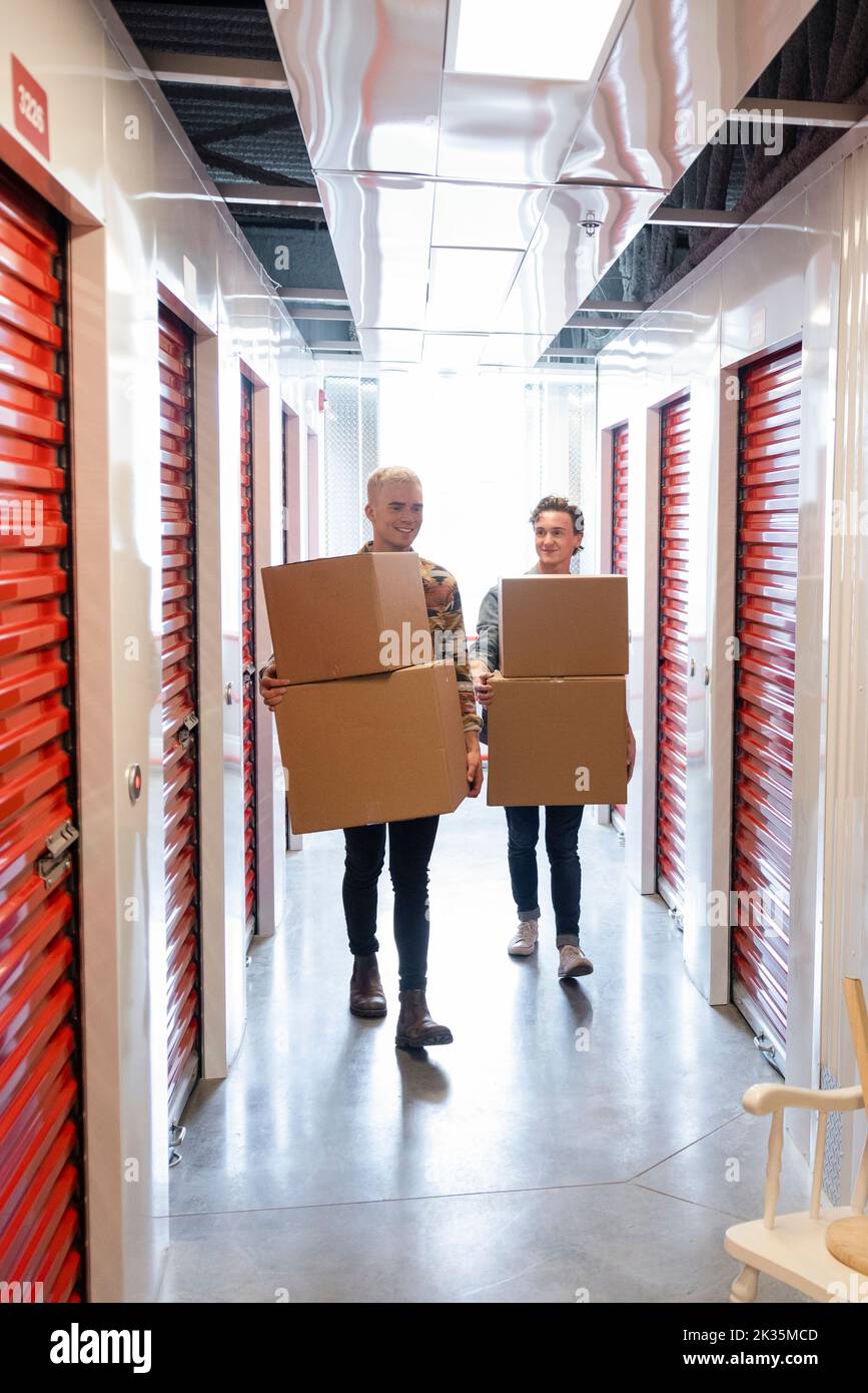 Young men carrying cardboard boxes in storage facility corridor Stock