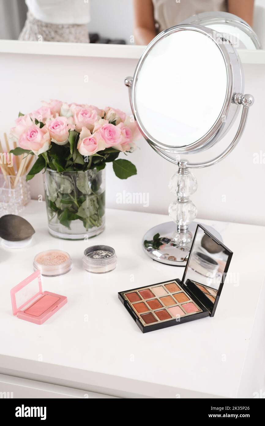 Cosmetics for make-up, round mirror and roses flowers on table. Decorative cosmetics, brushes, eyes shadow with foundation on dressing table in beauty Stock Photo