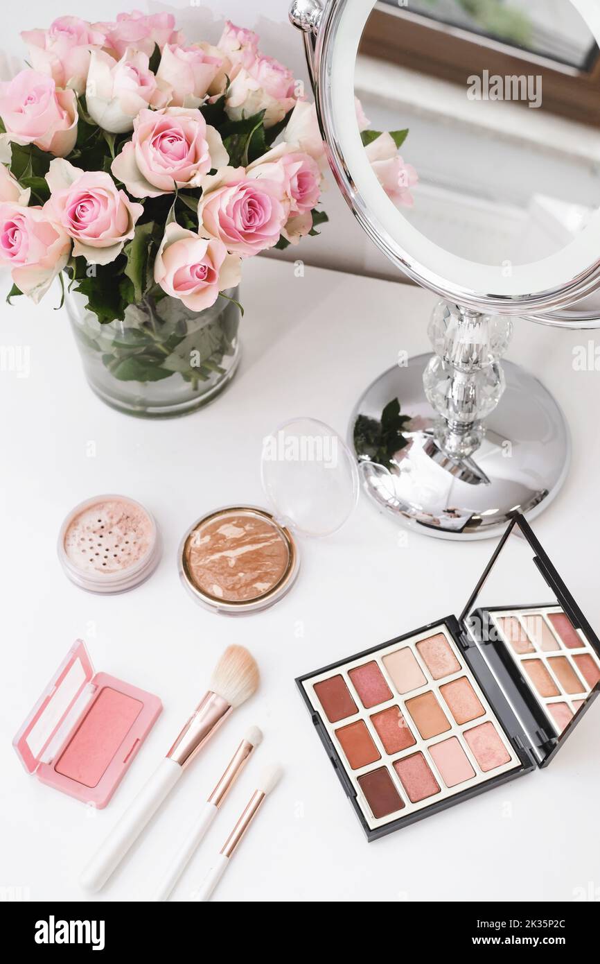Cosmetics for make-up, round mirror and roses flowers on table. Decorative cosmetics, brushes, eyes shadow with foundation on dressing table in beauty Stock Photo