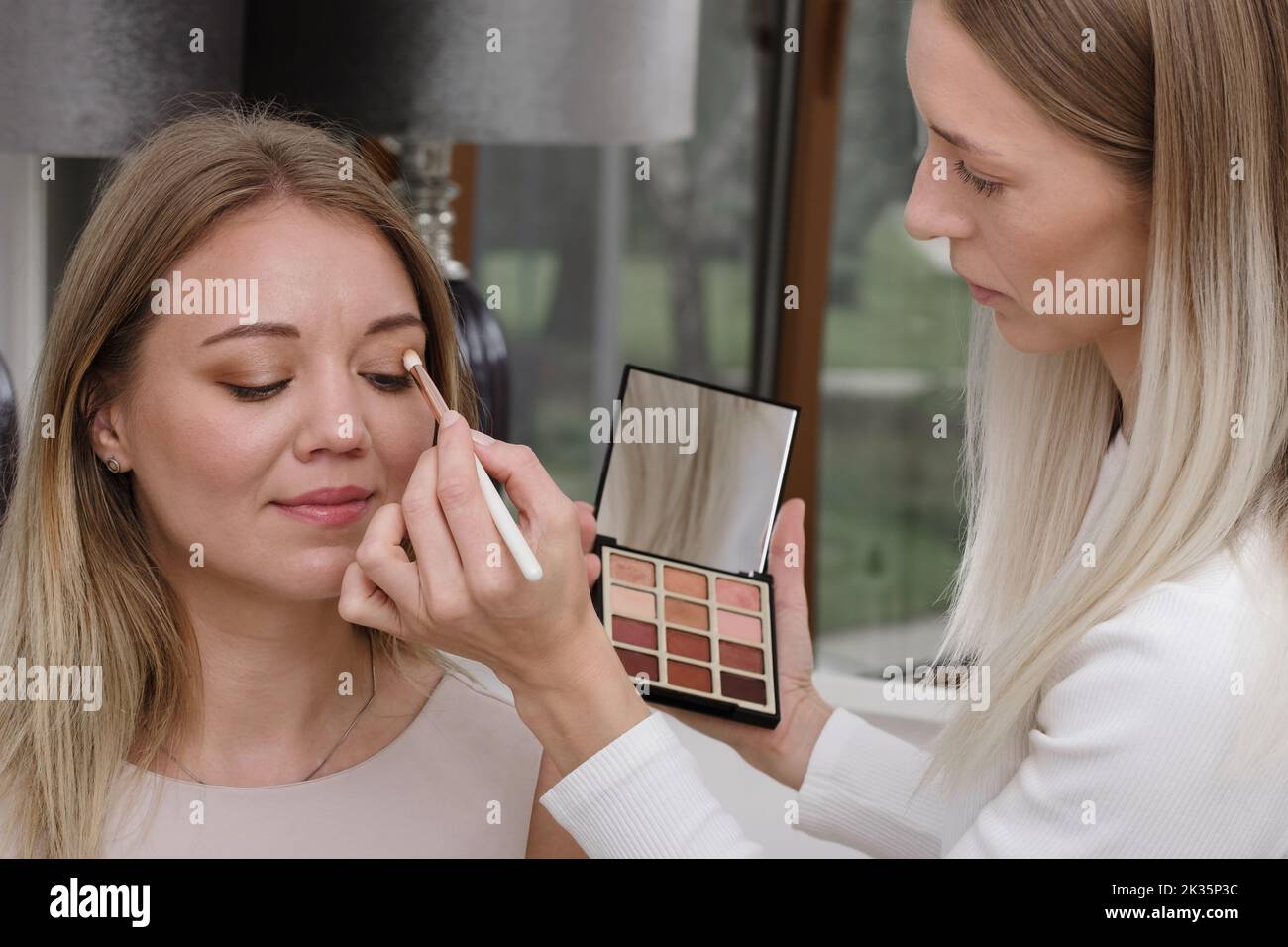 Make-up artist working with client in light beauty salon. Woman applying decorative cosmetics in front of mirror for blond hair model. Beautician Stock Photo