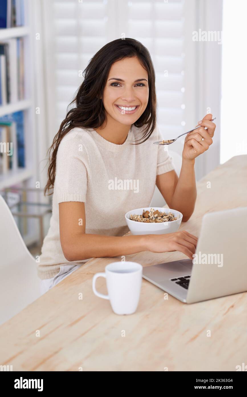 Technology at the breakfast table. Portrait of an attractive young woman using her laptop at home while eating breakfast. Stock Photo