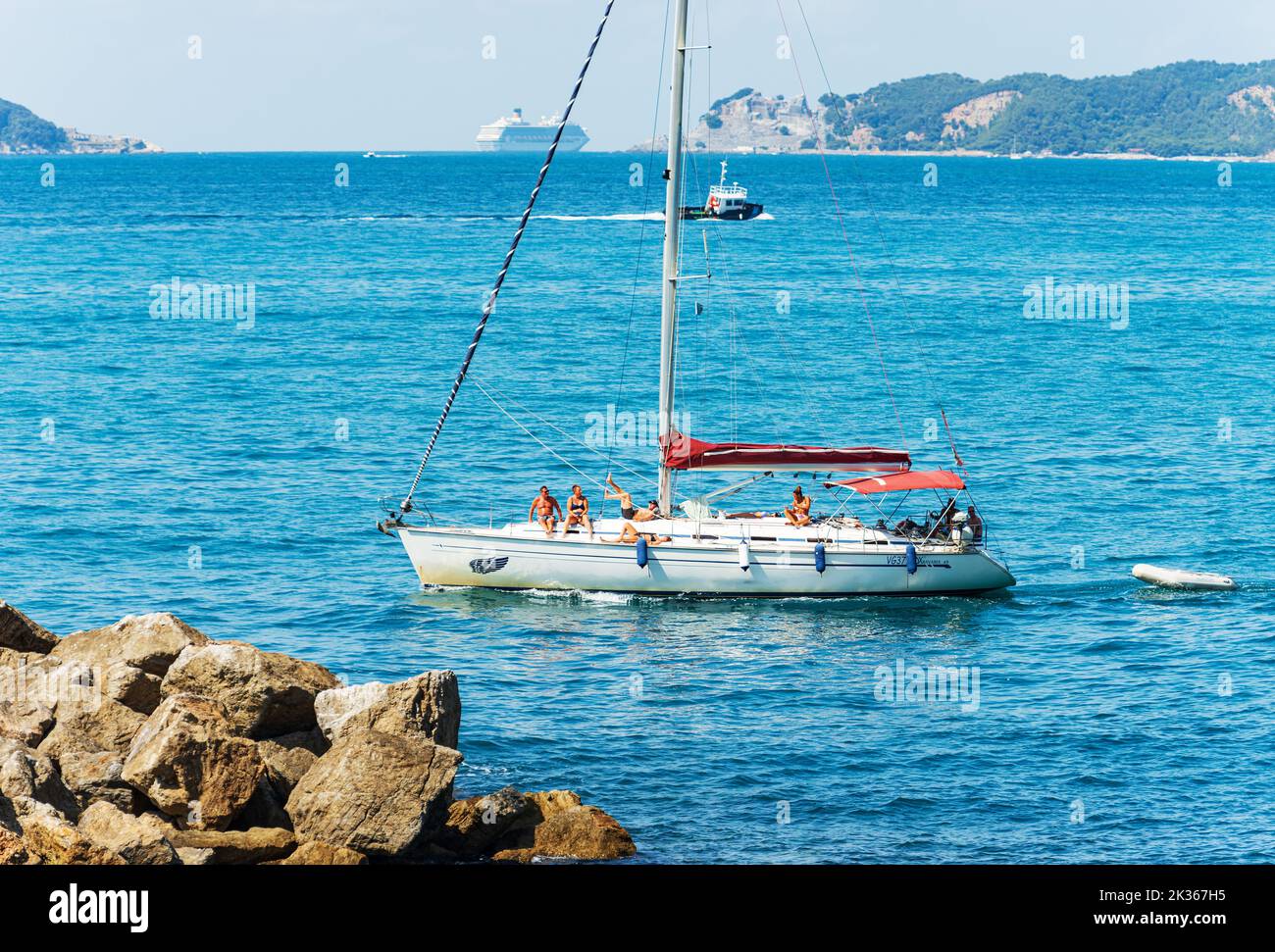 White sailboat with a group of people on board and a cruise ship, Tellaro village, Lerici, Mediterranean Sea, Gulf of La Spezia, Liguria, Italy. Stock Photo