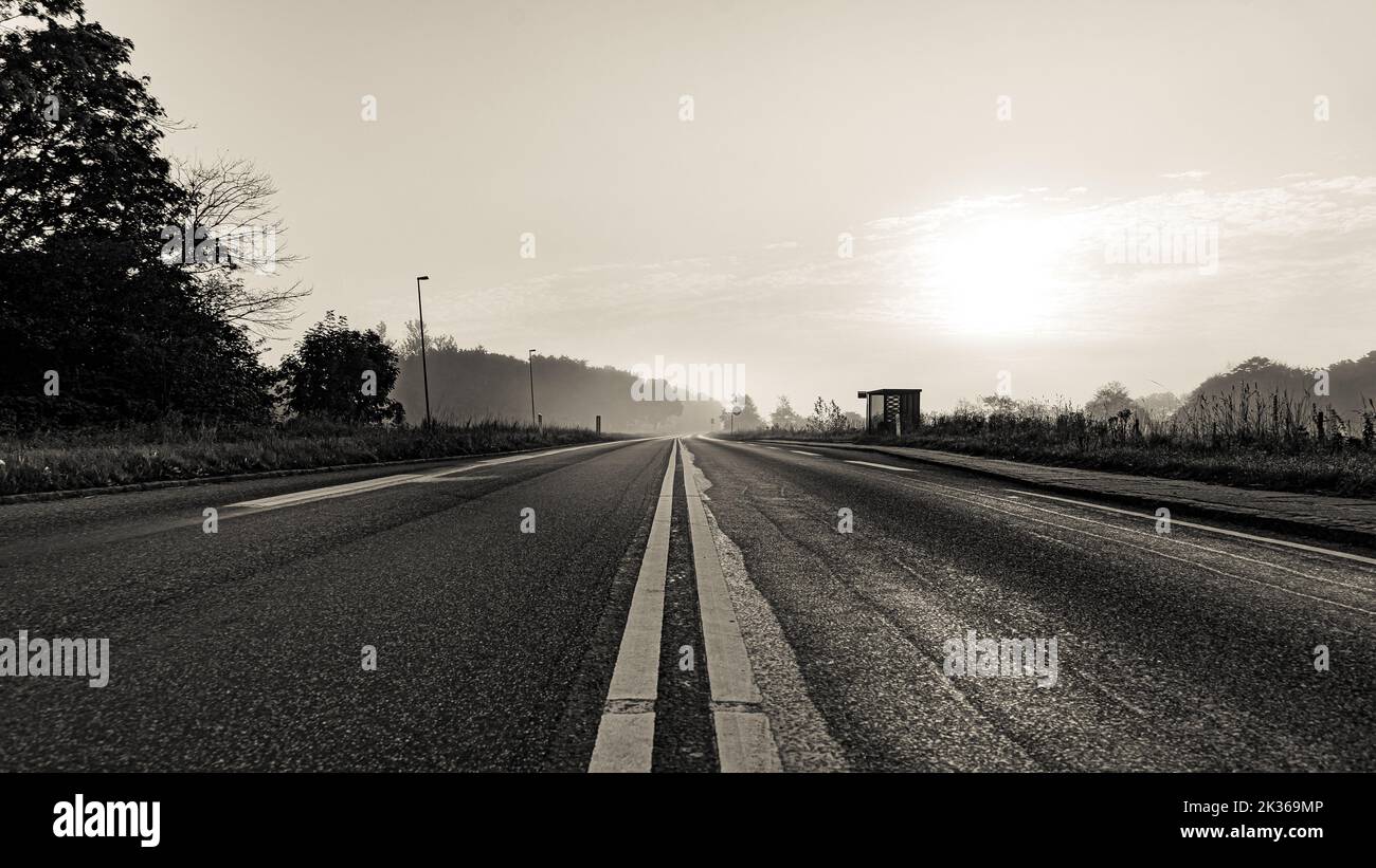 Black-and-white photograph of a rural road with a lonely bus stop in the early morning light Stock Photo