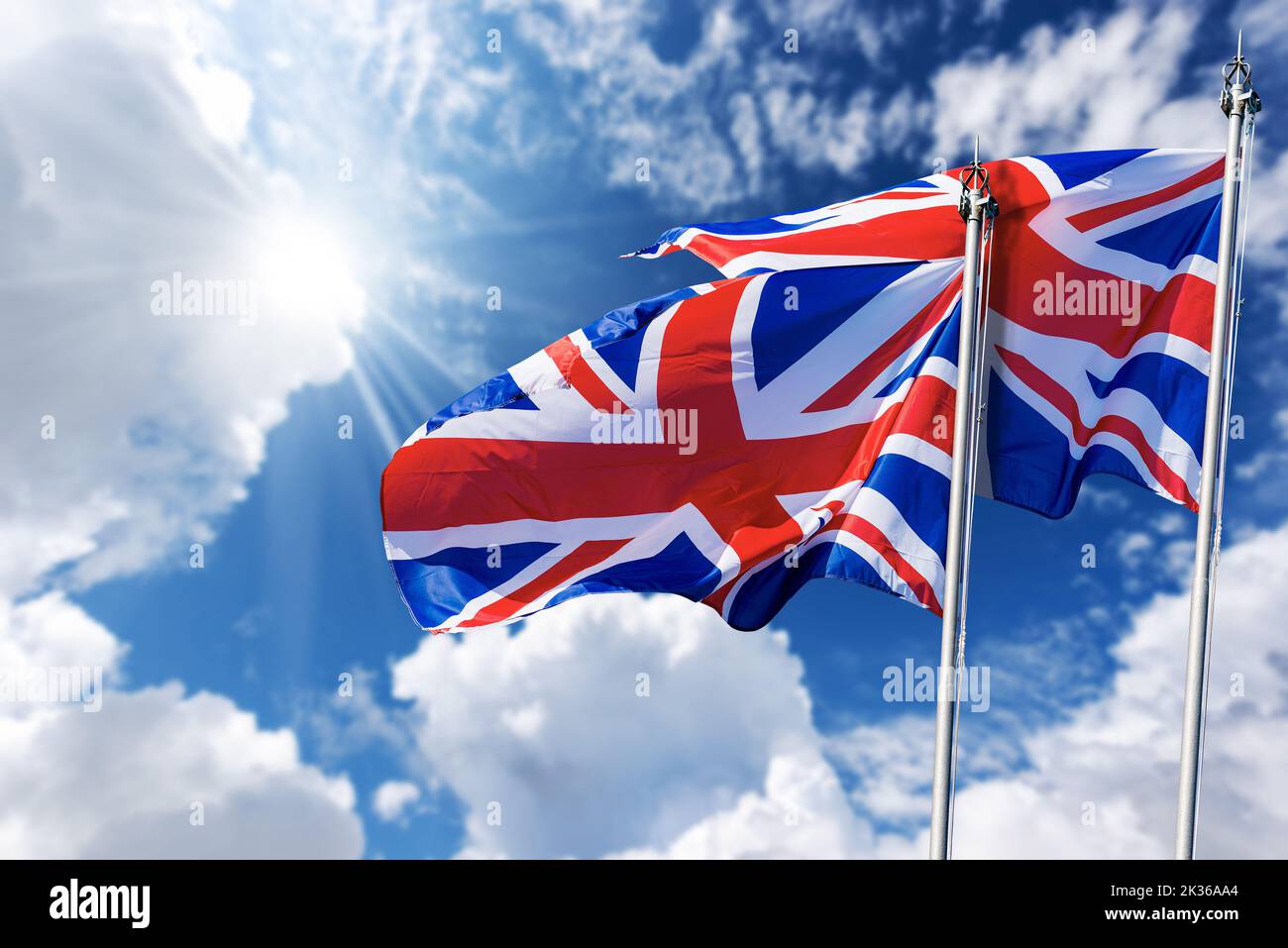 Two national UK flags with flagpole, blowing in the wind on a blue sky with clouds and copy space and sunbeams. Union Jack Flag. Stock Photo
