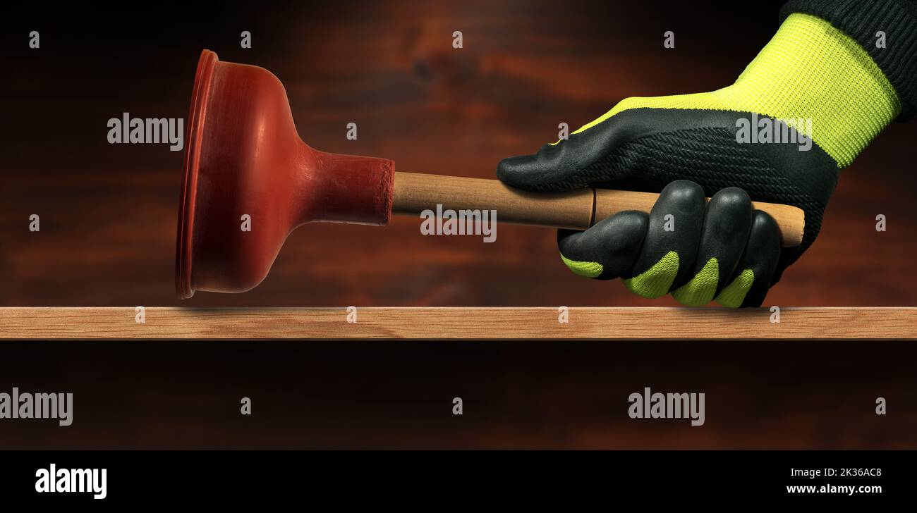 Hand with green and black protective work glove, holding a red rubber plunger with wooden handle, on a wooden workbench with copy space. Stock Photo