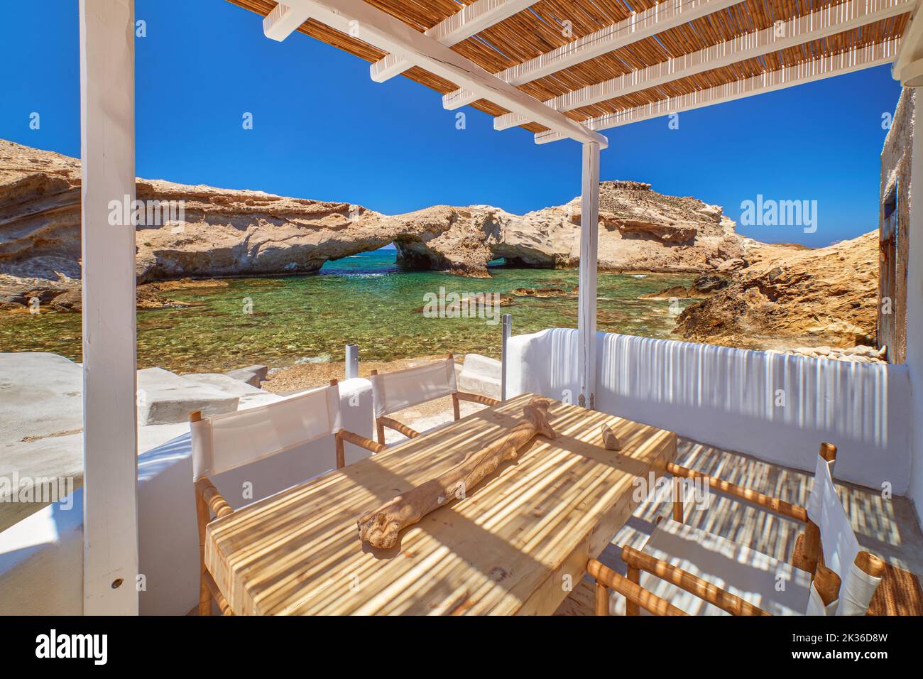 Terrace of summer villa by seafront on sunny day. Small bay, no people, clear sea waters, clear blue sky, sunshine, rocks and caves. Milos, Greece Stock Photo