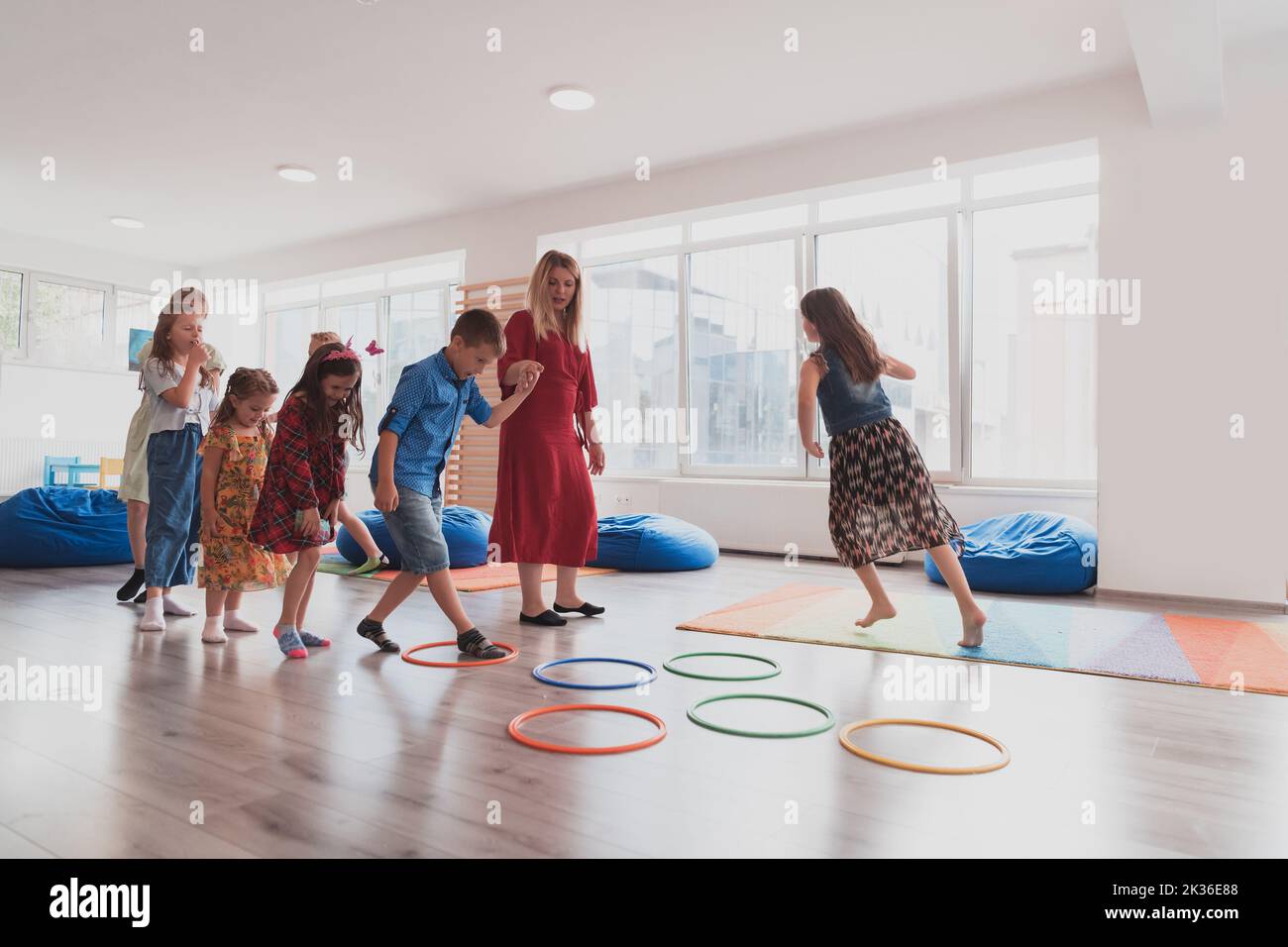 Small nursery school children with female teacher on floor indoors in classroom, doing exercise. Jumping over hula hoop circles track on the floor. Stock Photo