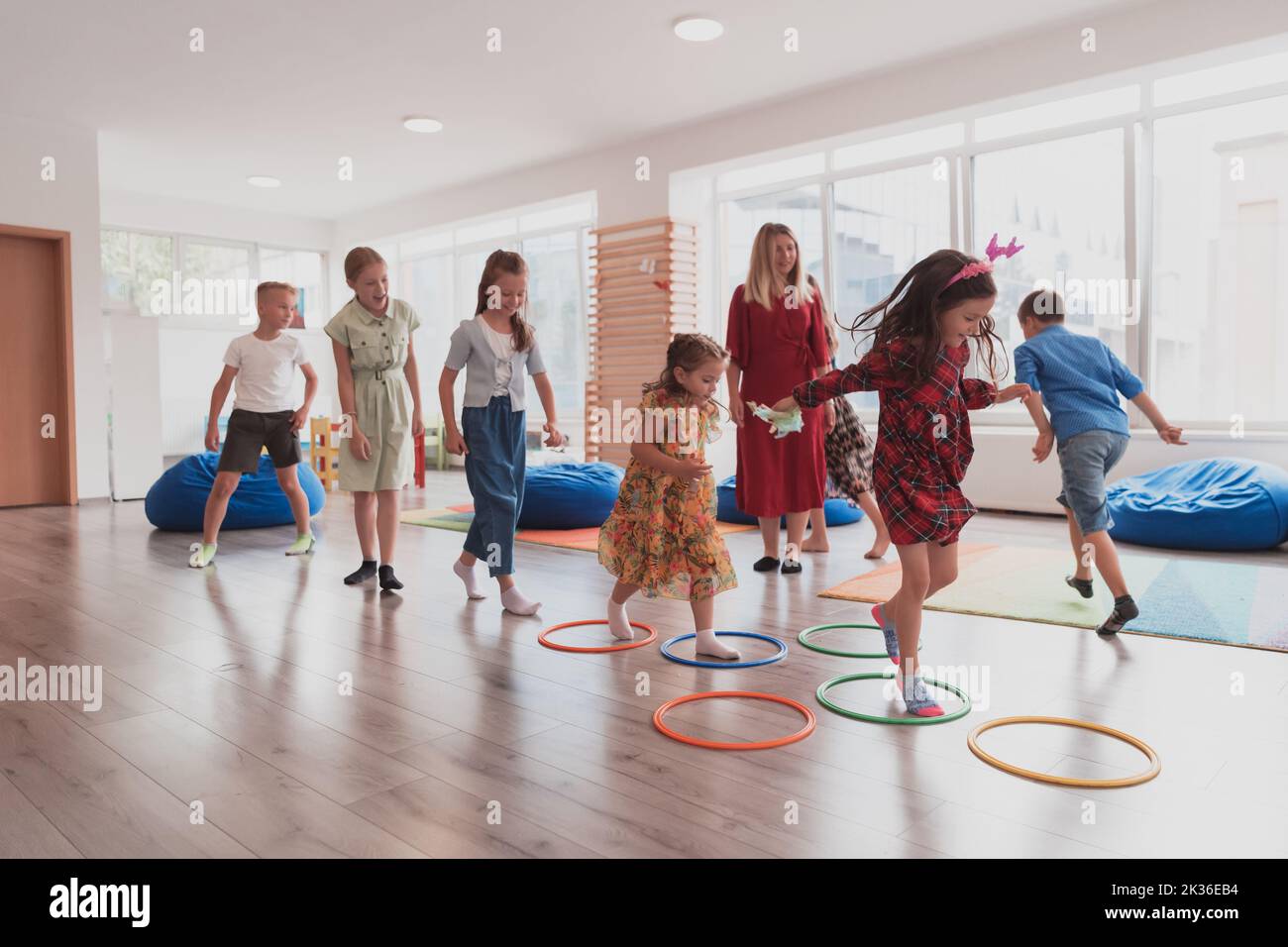 Small nursery school children with female teacher on floor indoors in classroom, doing exercise. Jumping over hula hoop circles track on the floor. Stock Photo