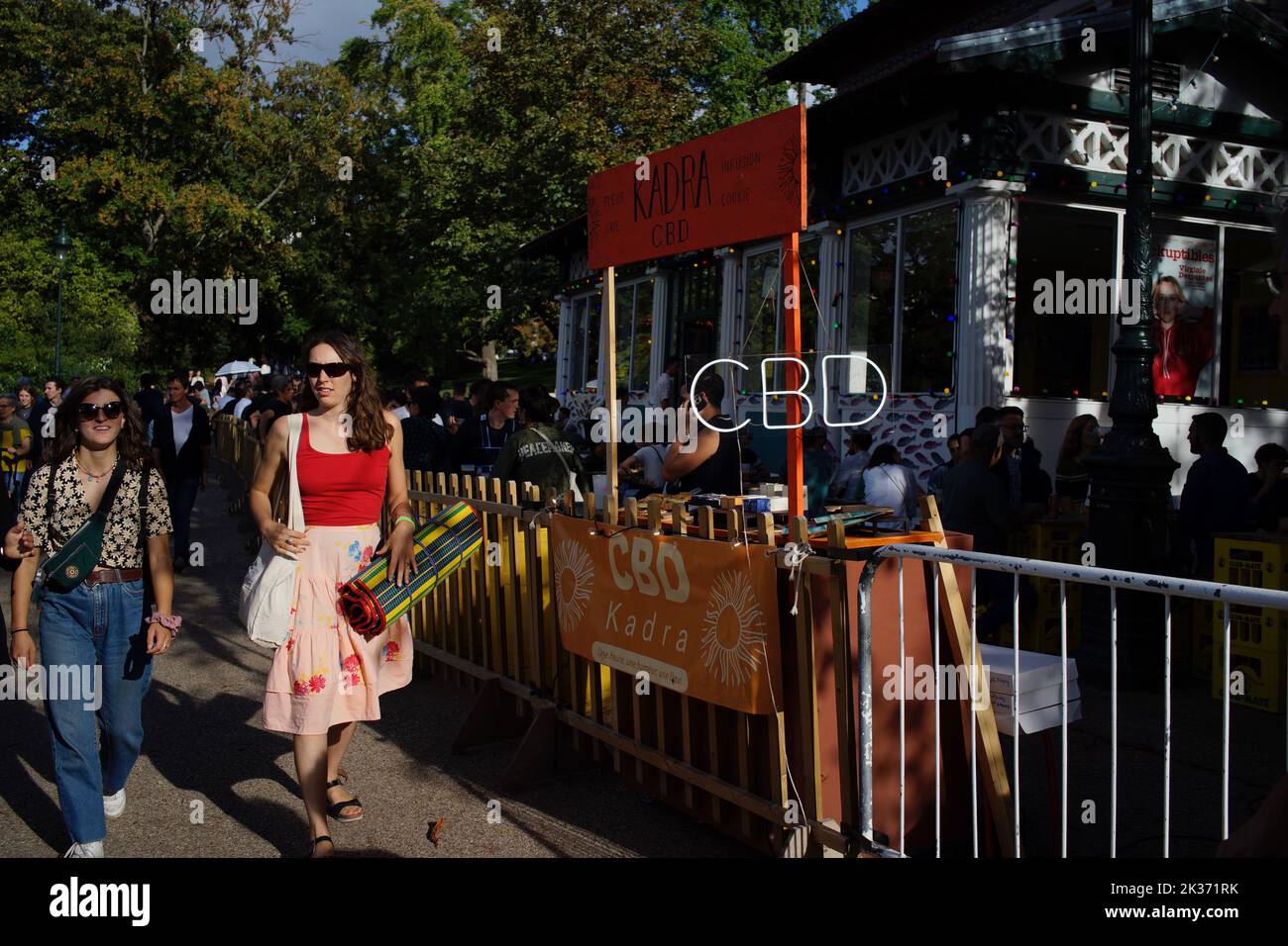 Woman walk by a 'Kadra' CBD stall selling products containing Cannabidiol, outside a busy Rosa Bonheur café, Parc des Buttes Chaumont, Paris, France Stock Photo