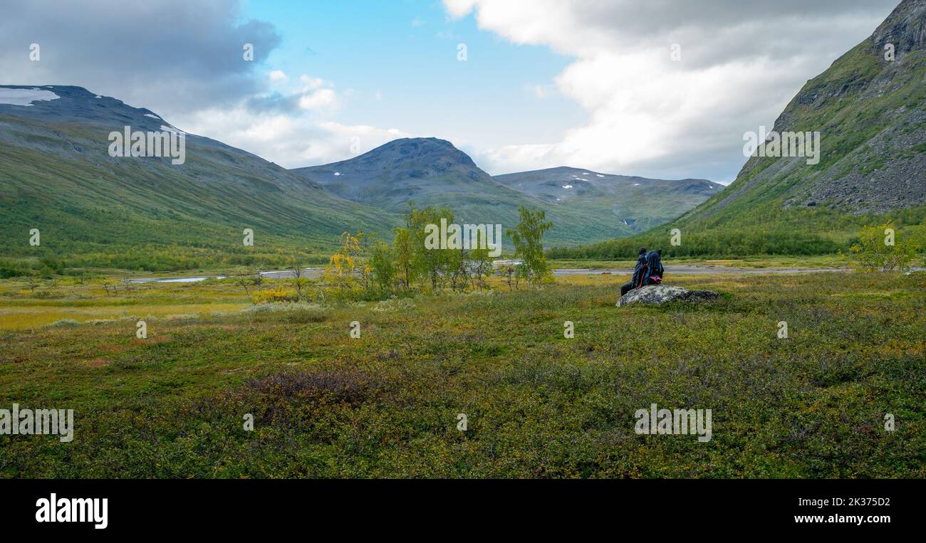 Female hiker with heavy gear sitting on a boulder, overlooking vast Arctic valley Rapadalen in Sarek National Park, Lapland, Sweden. Hiking day in Stock Photo