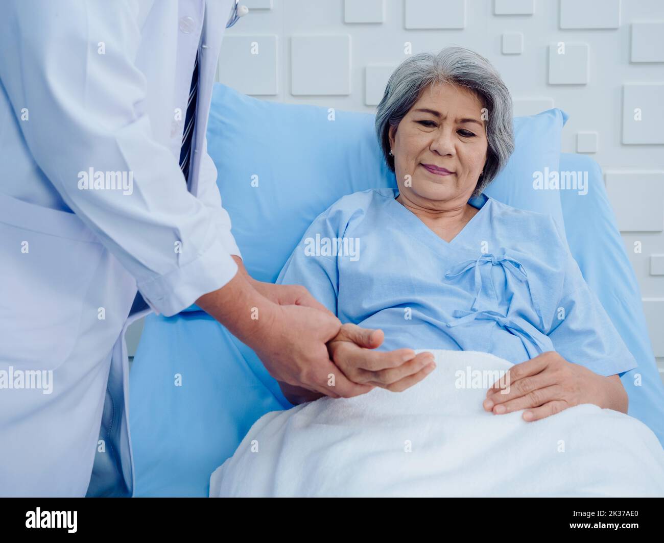 Happy Asian elderly patient woman smiling on bed while doctor in white lab coat holding hand for check to examine heartbeat pulse in recovery room in Stock Photo